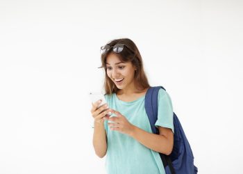 Portrait of brown haired Latin student girl in casual wear carrying backpack on one shoulder having shocked look, reading astonishing news on web site using 3G internet connection on mobile phone