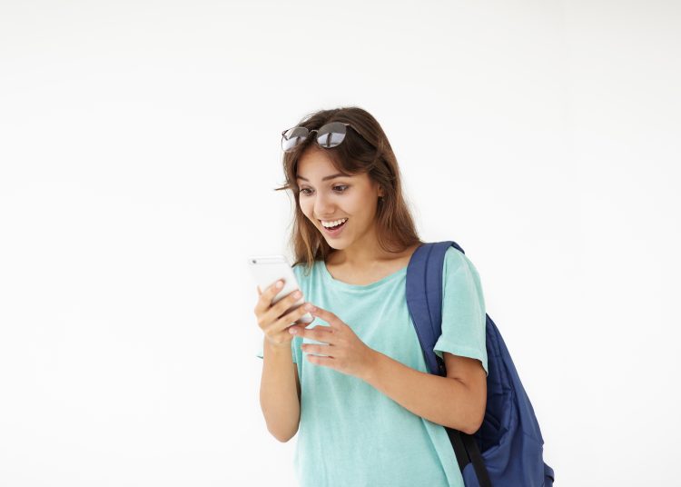 Portrait of brown haired Latin student girl in casual wear carrying backpack on one shoulder having shocked look, reading astonishing news on web site using 3G internet connection on mobile phone