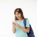 Portrait of brown haired Latin student girl in casual wear carrying backpack on one shoulder having shocked look, reading astonishing news on web site using 3G internet connection on mobile phone