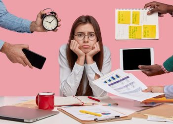 sad-female-workaholic-keeps-hands-chin-busy-making-project-work-studies-papers-wears-elegant-white-shirt-sits-desktop-unknown-people-stretch-hands-with-notes-alarm-clock-smartphone_273609-26174