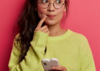 photo-pensive-surprised-woman-keeps-index-finger-near-lips-looks-aside-thinks-about-message-content-holds-mobile-phone_273609-39036